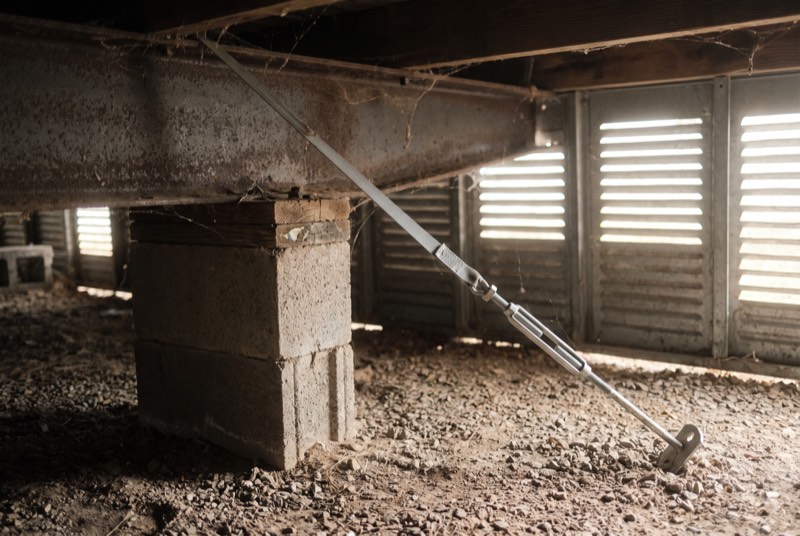 A steel chassis beam on a concrete block pier with a tie-down strap under a manufactured home